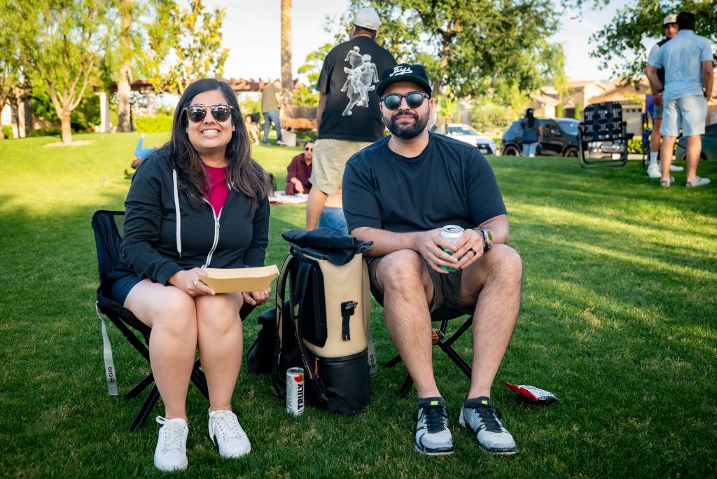 Two people sit on lawn chairs in a park, smiling at the camera. One holds food, the other a drink. They wear sunglasses and casual clothes. Other people and trees are visible in the background on a sunny day.