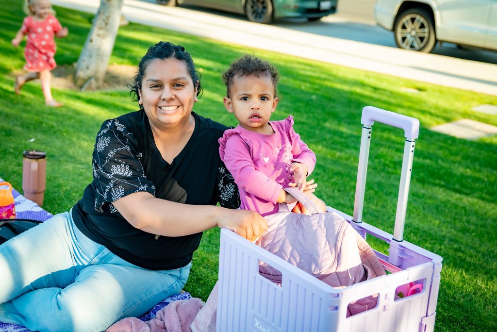 A woman sits on the grass smiling while a young child in pink stands beside her, holding onto a purple crate. They are outdoors on a sunny day, with cars and another child in the background.