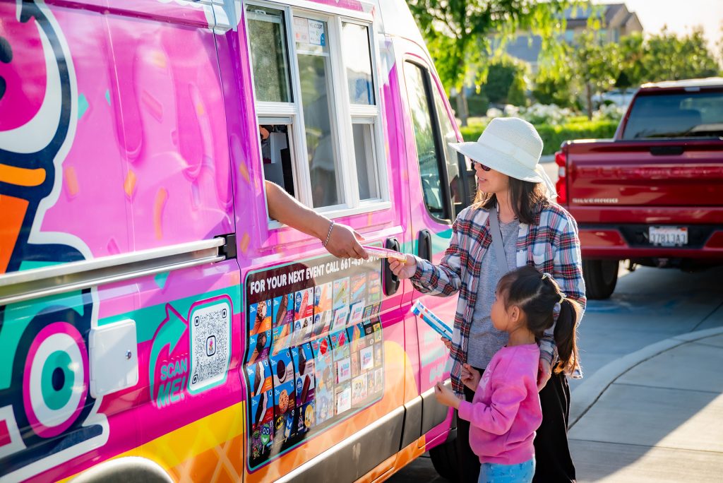 A woman and a young girl buy ice cream from a colorful ice cream truck parked on a sunny street. The woman hands cash to the vendor through the window while the girl holds a treat and smiles.