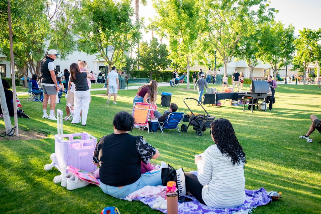 People gather in a sunny park, sitting on blankets and lawn chairs. Some stand and chat while others set up coolers and strollers. Trees provide shade and the atmosphere is relaxed and social.