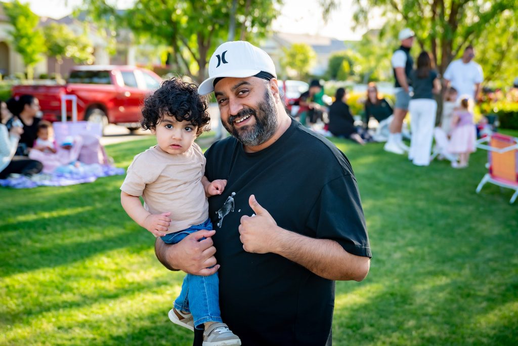 A man wearing a white cap and black shirt smiles and gives a thumbs up while holding a young child on a grassy lawn, with families and children gathered in the sunny background.