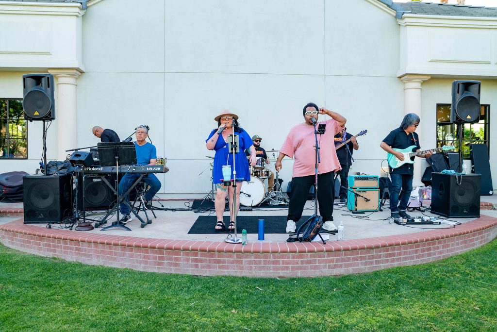 A band performs outdoors on a small stage. There are six members: a keyboardist, drummer, two vocalists (one in a blue dress and hat, one in a pink shirt), a guitarist, and a bassist. Equipment and speakers surround them.