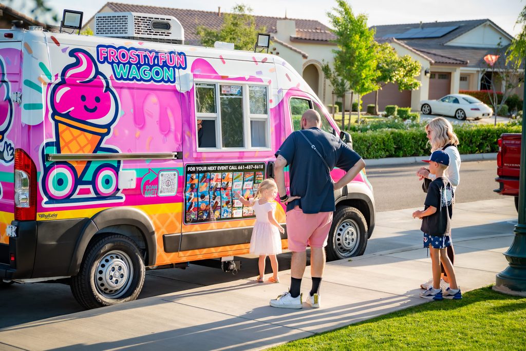A family with children stands at a colorful ice cream truck labeled "Frosty Fun Wagon" on a sunny suburban street, choosing treats from the menu on the side of the vehicle.