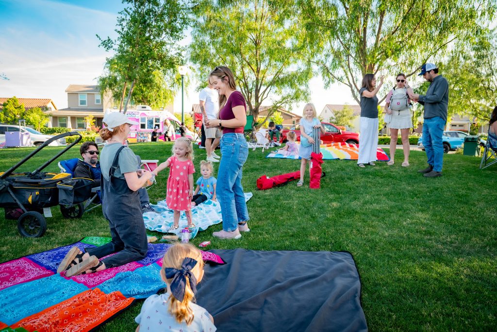 Families and children gather on a grassy lawn for a picnic, with colorful blankets and trees around. Some adults stand and chat while kids play nearby. Houses and a pink food truck are visible in the background.