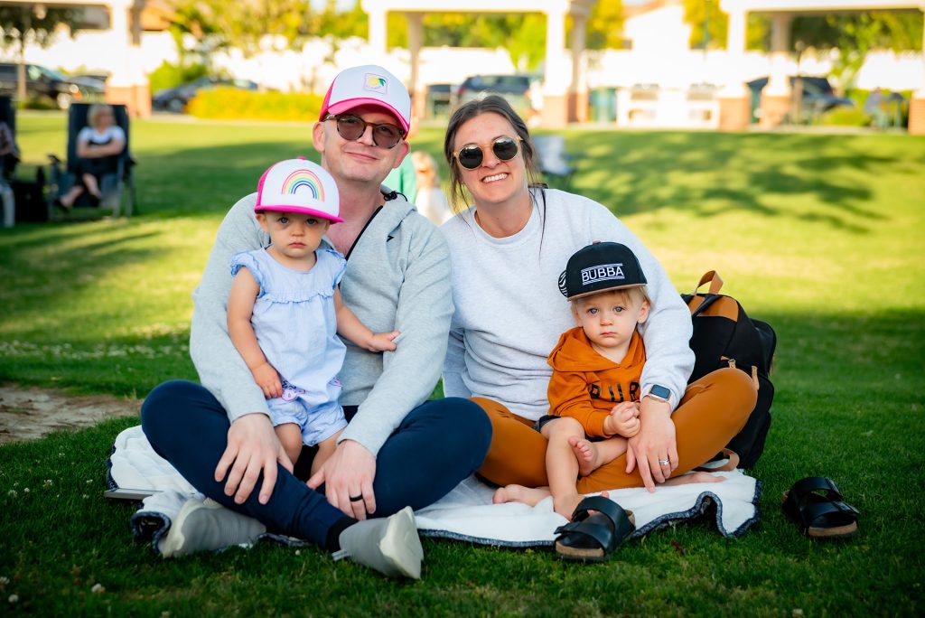 A family of four sits on a blanket outdoors. Two adults sit in the middle, each holding a young child on their lap. They all wear casual clothes and hats, smiling and enjoying a sunny day at the park.