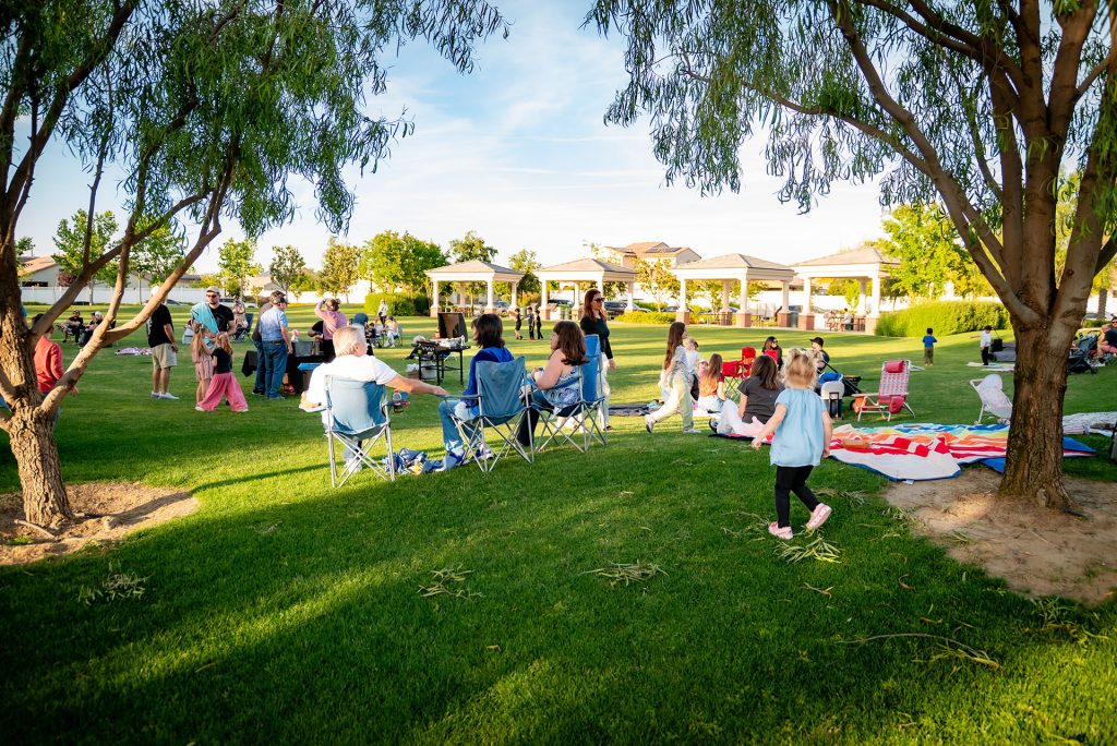 People gather outdoors in a park, sitting on folding chairs and blankets under trees. Children play on the grass, and others stand near picnic tables. Pavilions and houses are visible in the background on a sunny day.