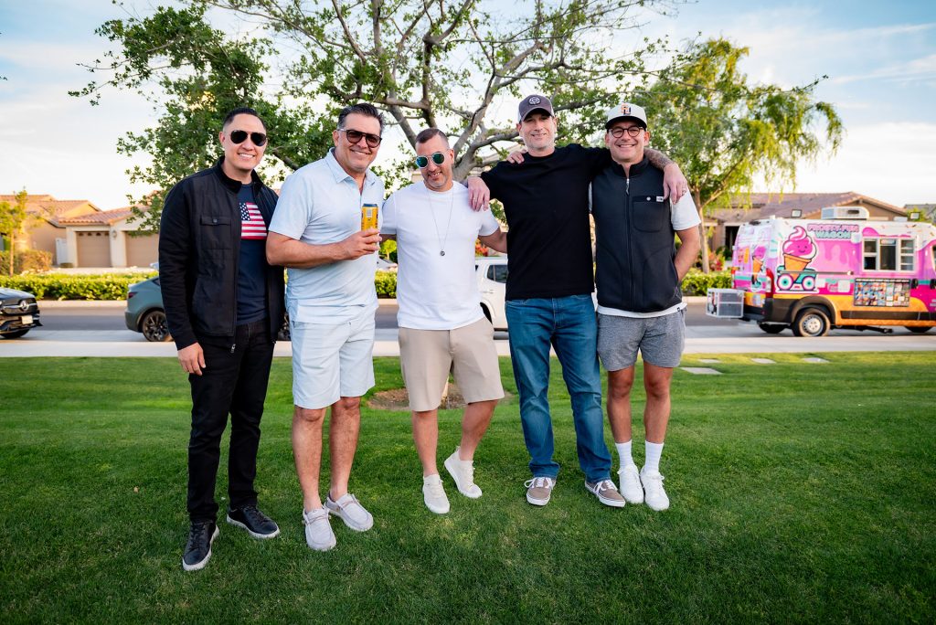 Five men stand together smiling on a grassy lawn, with an ice cream truck and houses in the background on a sunny day. They are casually dressed and appear to be enjoying a relaxed outdoor gathering.