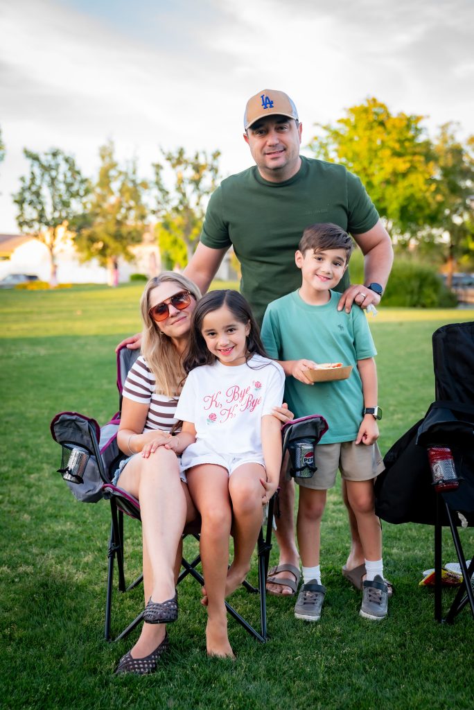 A smiling family of four poses outdoors on grass. The mother, in sunglasses, sits on a chair with a young girl in her lap. The father stands behind them with a young boy, both smiling. Trees and sunlight fill the background.