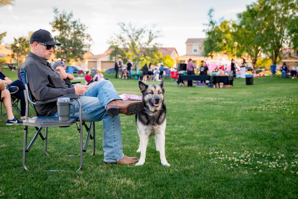 A man wearing sunglasses sits on a lawn chair with a large dog beside him on a grassy field. People and tents are visible in the background, suggesting a community outdoor event.
