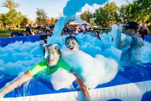 Two children laugh and play in a large foam-filled pool at an outdoor event, surrounded by other kids and bubbles, with trees and people in the background under a bright sky.