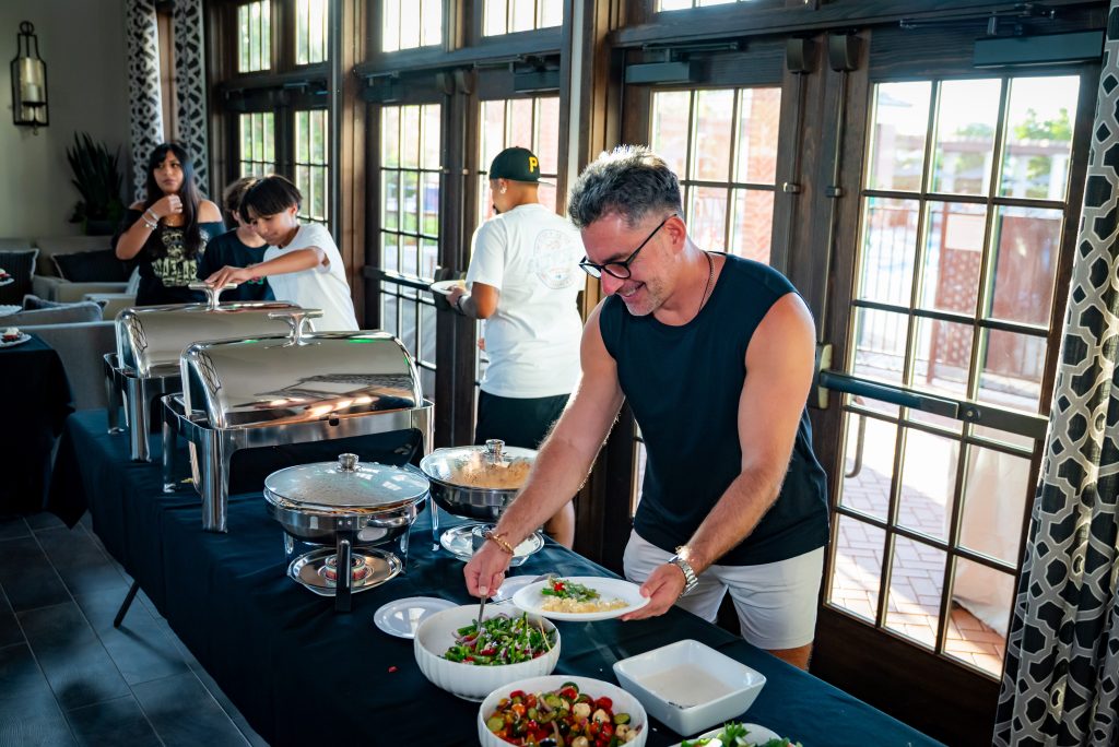 A man in a sleeveless shirt serves himself food from a buffet table with salads and covered dishes, while others, including children, gather food in a bright room with large windows.