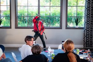 A performer in a red jacket and sunglasses strikes a dynamic pose with a microphone, entertaining an audience seated at tables inside a bright room with large windows and plants. Audience members film the performance on their phones.