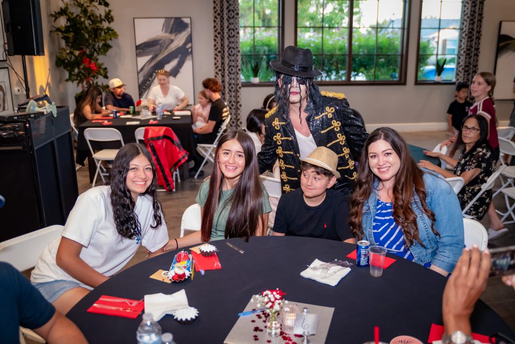 A group of four smiling kids sits at a round table with party decorations, while a person dressed as Michael Jackson stands behind them. Other people are seated at tables in the background of the brightly lit room.