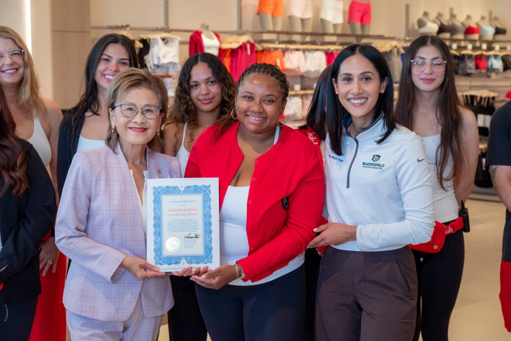 A group of women smile together indoors as one woman presents a certificate of appreciation to another, with others gathered around them in athletic wear.