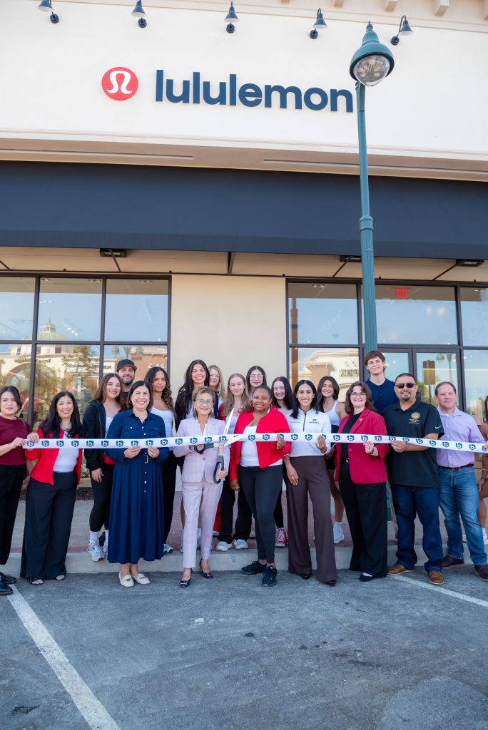 A group of people pose in front of a Lululemon store for a ribbon-cutting ceremony, holding the ribbon together and smiling at the camera.