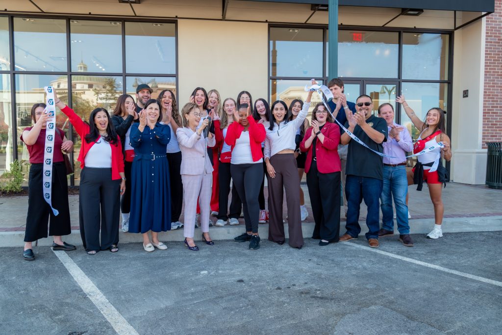 A group of people stand outside a building, smiling and cheering, while one woman holds a long ribbon. Some are clapping and a few wear red jackets. It appears to be a celebratory event, possibly a ribbon-cutting ceremony.