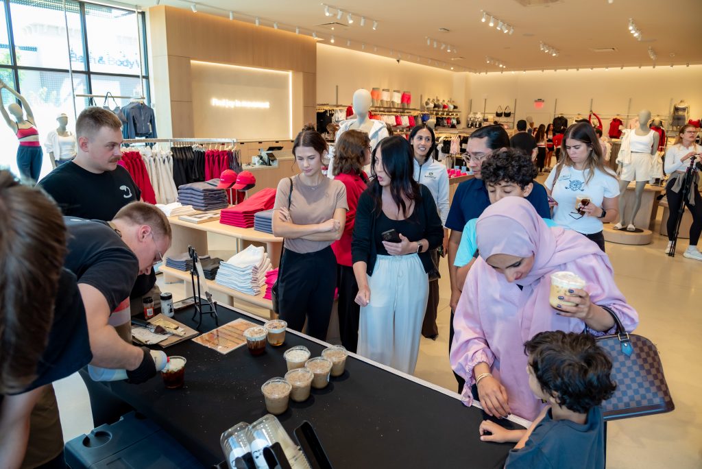 People crowd around a table with drinks at a clothing store event. Some adults and children watch as drinks are prepared. Mannequins and folded clothes are visible in the bright, modern store.