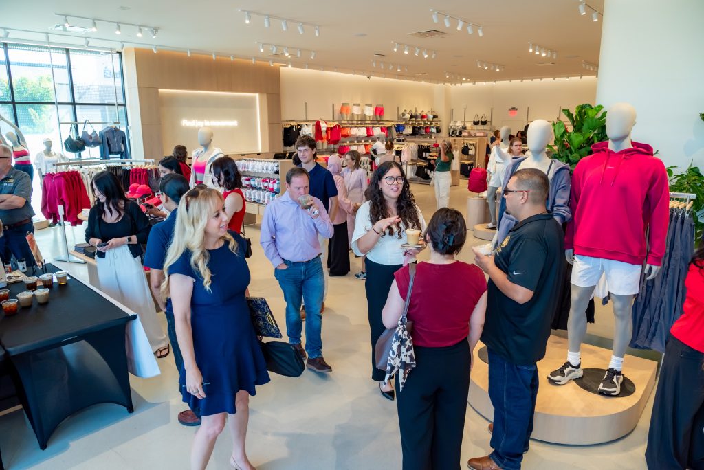 People socializing in a modern clothing store, with mannequins dressed in sportswear and racks of clothes visible. Bright lighting, casual atmosphere, and some people holding drinks.