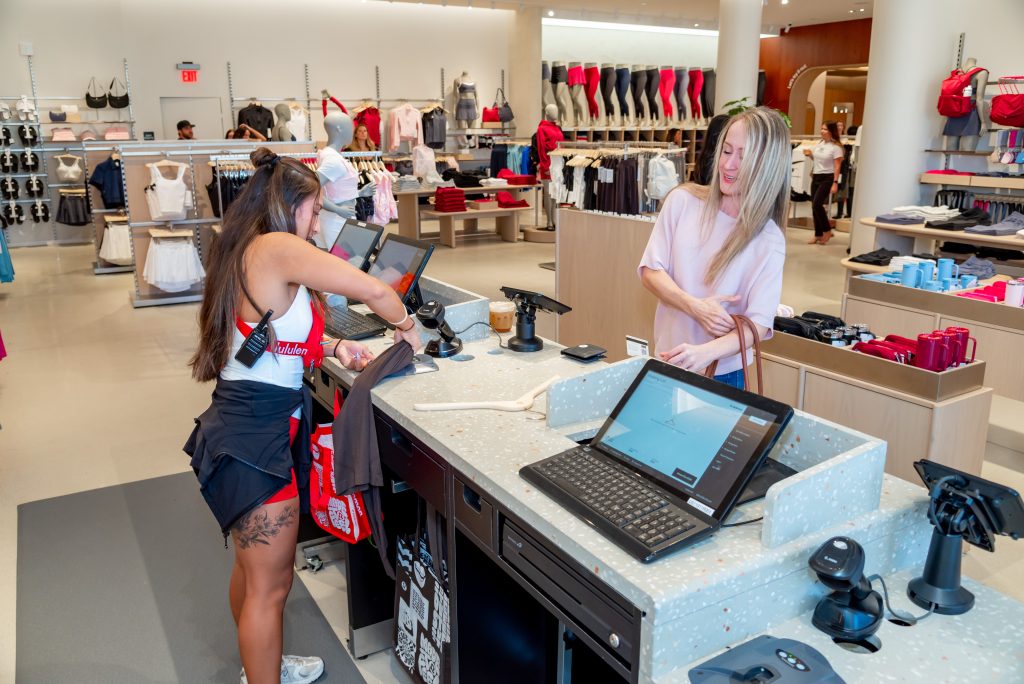 Two women at a retail store checkout counter, one staff member scans items while the other customer waits. Clothing displays and shopping bags are visible in the background.