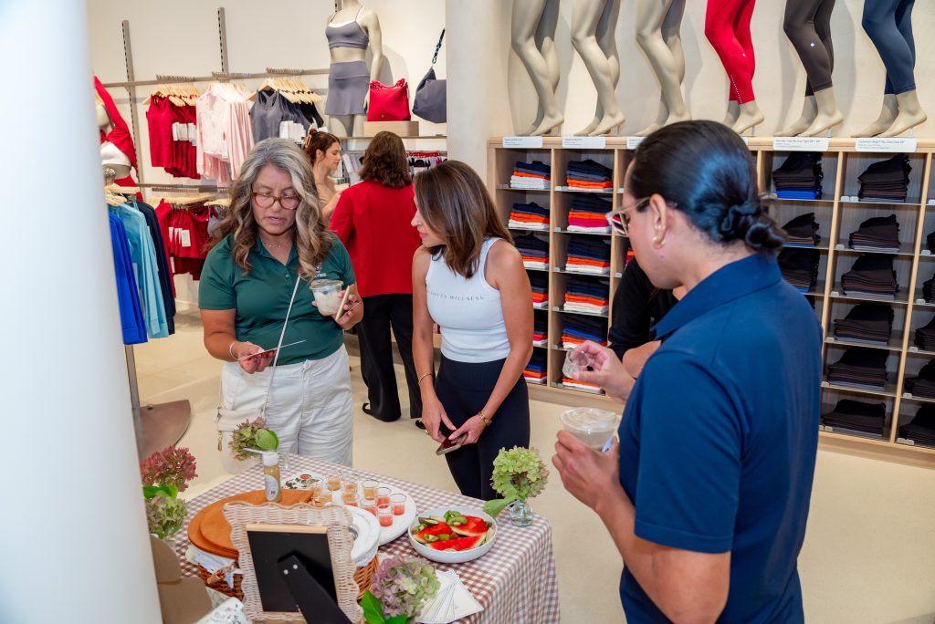 Four people stand around a small table with food samples in a clothing store, engaging in conversation. Shelves with folded activewear and mannequins in workout clothes are in the background.