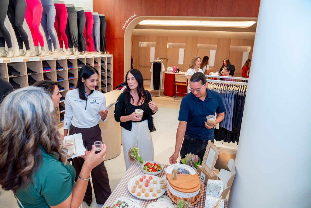 Four people stand around a table with drinks and food samples in a clothing store with racks of leggings and shelves in the background. Other shoppers are visible further inside the store.