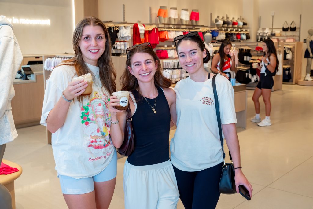 Three young women smile and hold drinks in a bright athletic apparel store, with shelves of clothes and accessories visible in the background. Other shoppers are browsing behind them.