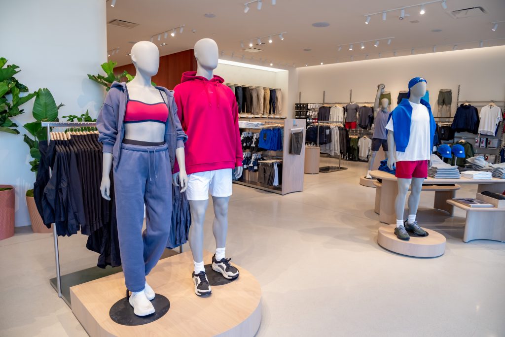 Three mannequins display athletic wear in a modern clothing store. Shelves and racks of sportswear and accessories are neatly arranged in the background, and large green plants decorate the space.