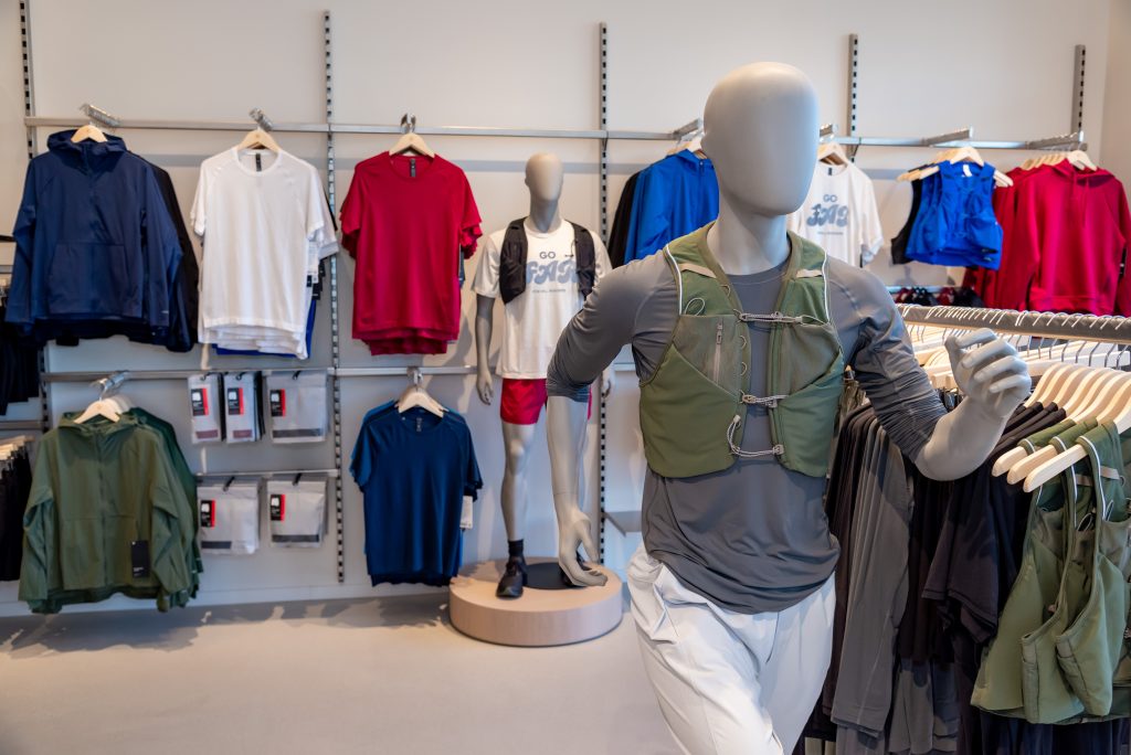 Mannequins in athletic wear are displayed in a store with shirts, jackets, and accessories hanging on racks and shelves in the background.