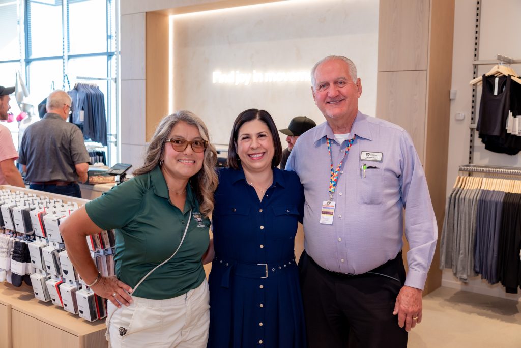 Three adults stand smiling inside a clothing store. Two women are on the left; a man is on the right wearing a work badge. Clothing items and displays are visible in the background.