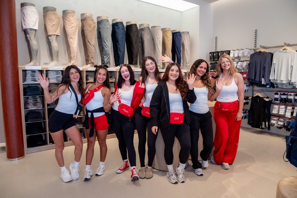 Seven women smile and wave in a clothing store, standing in front of a display of shorts and pants. They wear coordinated athletic outfits in red, white, and black.
