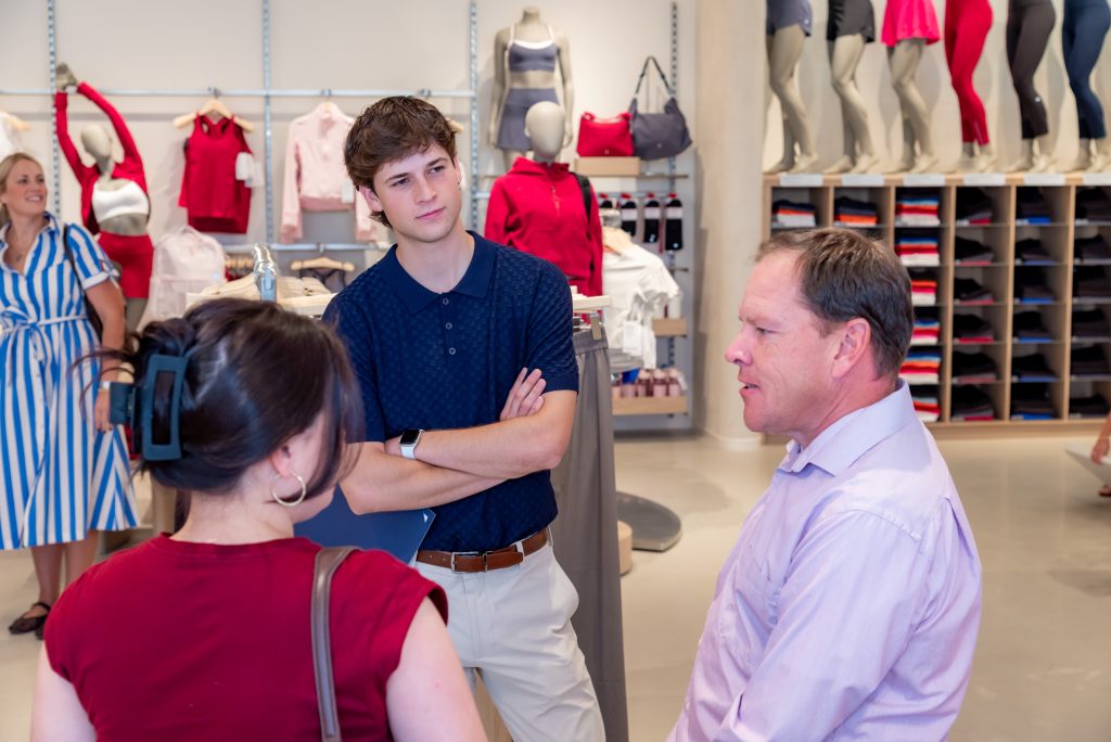 Three people are having a conversation inside a clothing store, with mannequins and folded clothes visible in the background. A woman in a blue and white dress stands further away, smiling.
