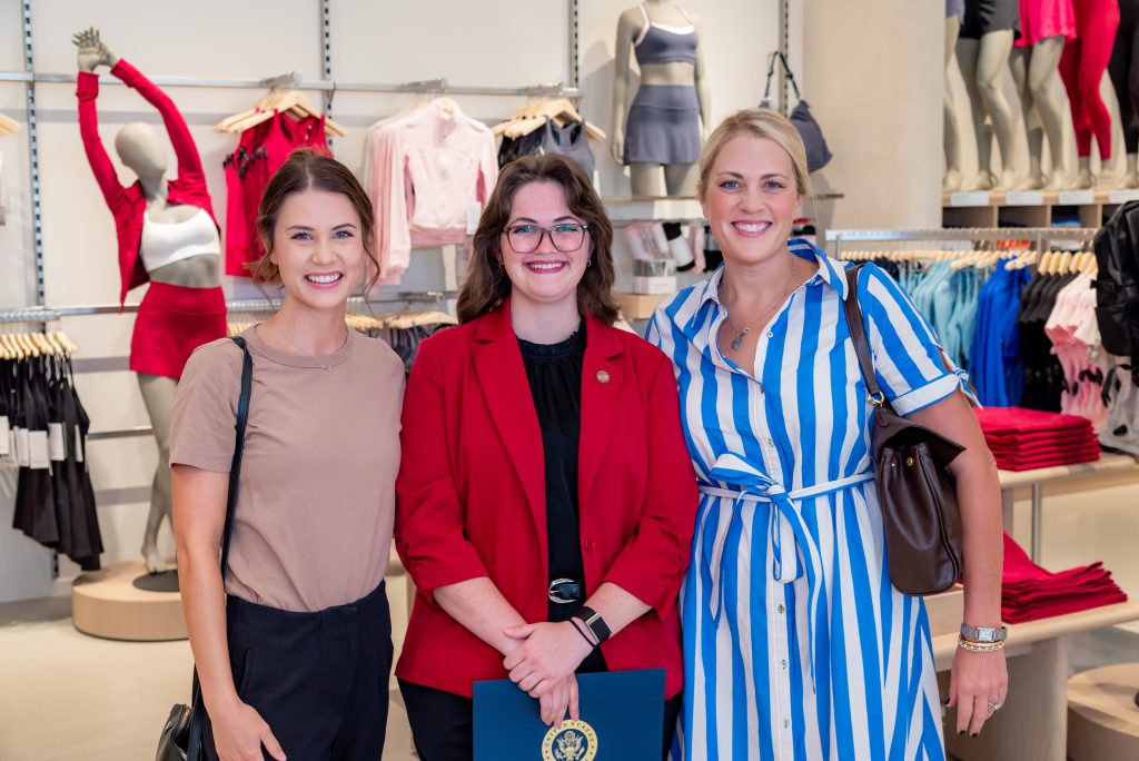 Three women smile together in a retail clothing store. Behind them are mannequins dressed in activewear and shelves with folded clothes. One woman holds a blue folder with a gold emblem.