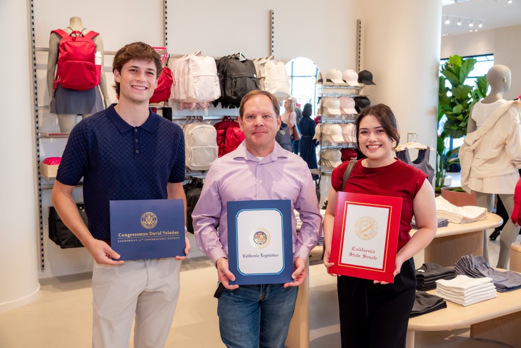 Three people stand in a clothing store, smiling and holding official certificate folders. Backpacks and shelves of clothes are displayed behind them.