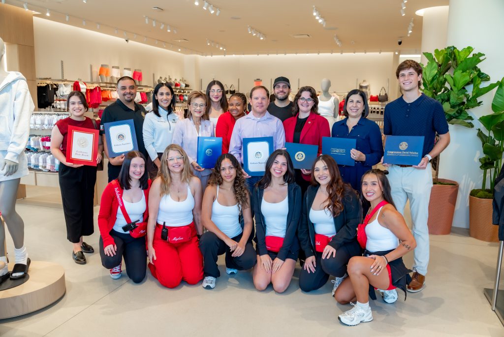 A group of 18 people pose inside a retail store, some kneeling and some standing, with several holding certificates and folders. Mannequins and clothing displays are visible in the background.