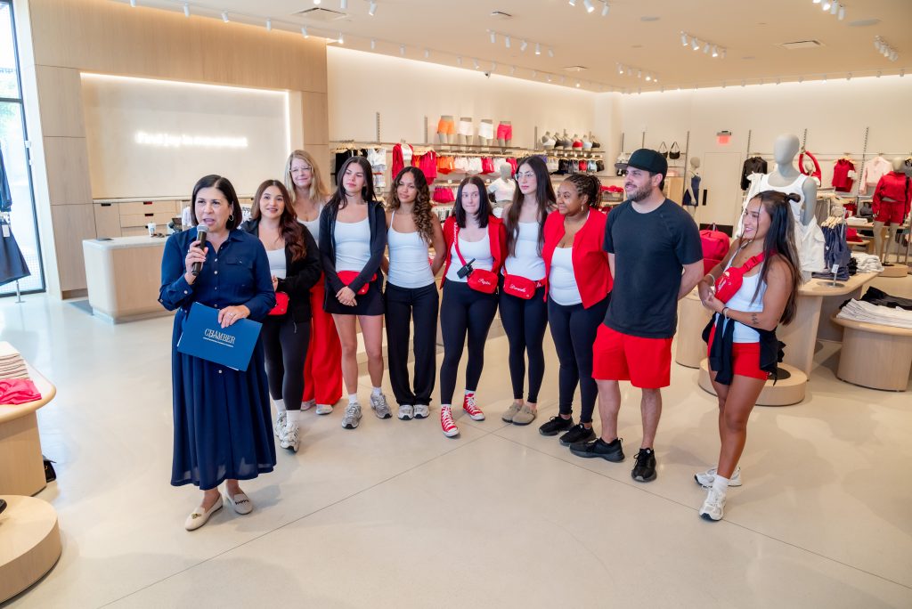 A woman speaks into a microphone while a group of people, mostly women in coordinated red and white outfits, pose together and smile inside a modern clothing store.