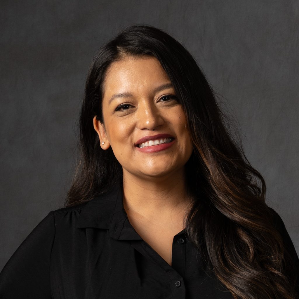 A woman with long dark hair and a black shirt smiles at the camera against a dark gray background.