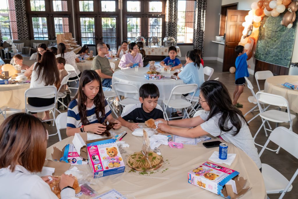 Children and adults sit around decorated tables at a party, assembling teddy bears from kits, with balloons and snacks visible in a brightly lit room.