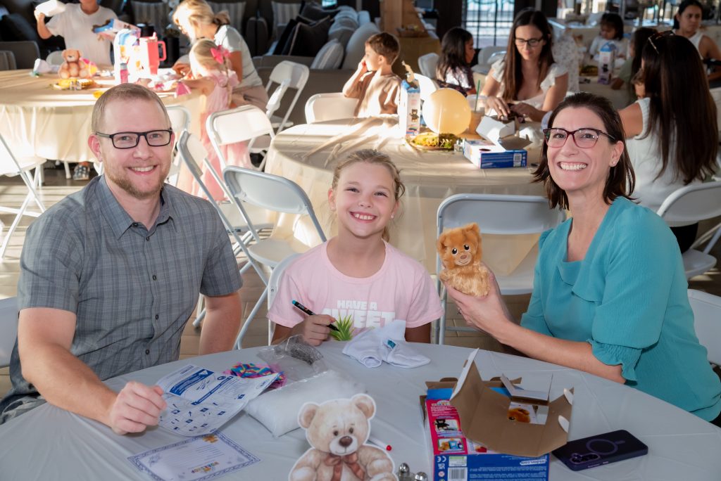 Three people sit at a table smiling, with a stuffed bear, papers, and a toy dog in front of them. Other groups are seen working at tables in a bright, busy room.