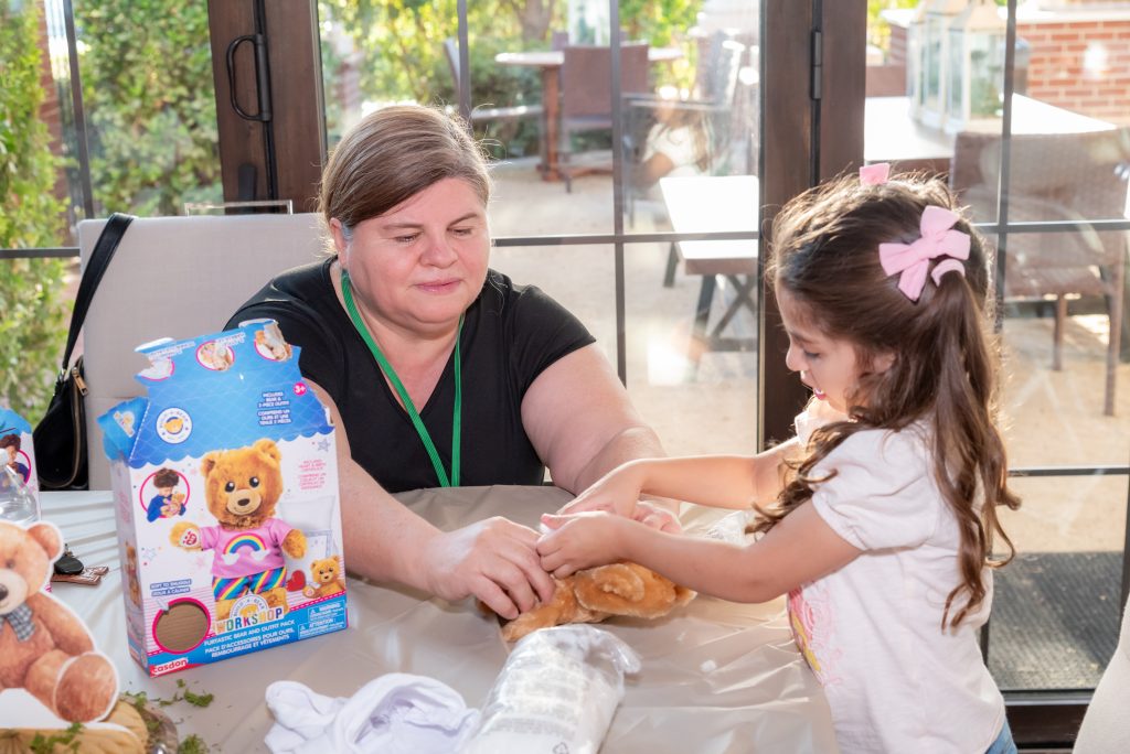 A woman and a young girl sit at a table making a teddy bear together. The table has a teddy bear kit, stuffing, and some white fabric. The girl is focused on the bear, while the woman watches and helps.