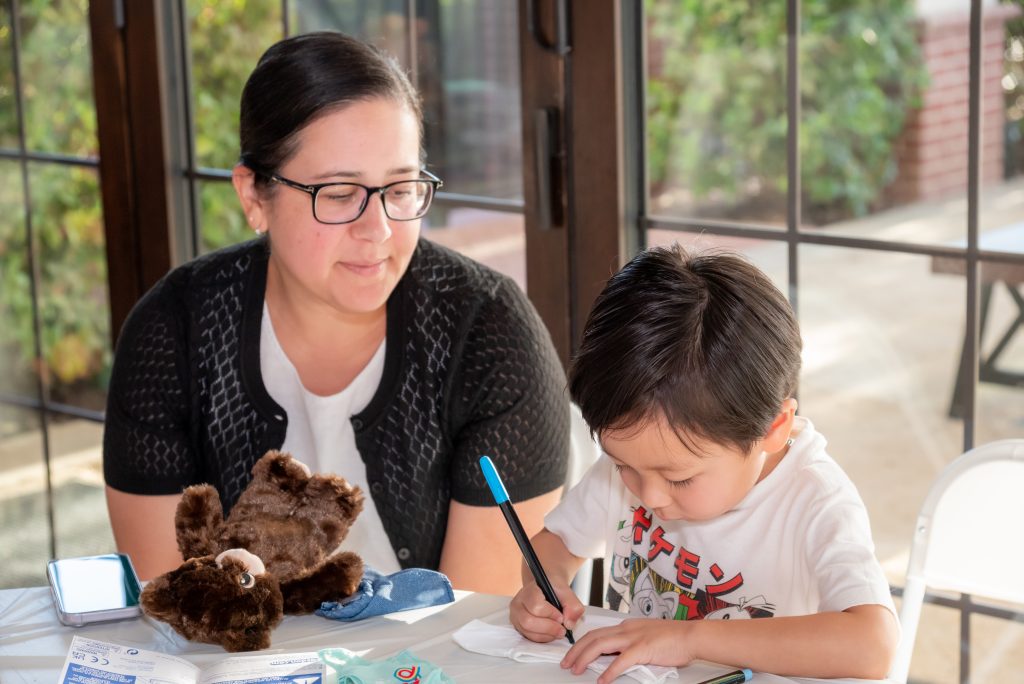 A woman watches a young boy drawing with a marker at a table. A plush teddy bear and art supplies are also on the table. Sunlight streams through large windows in the background.
