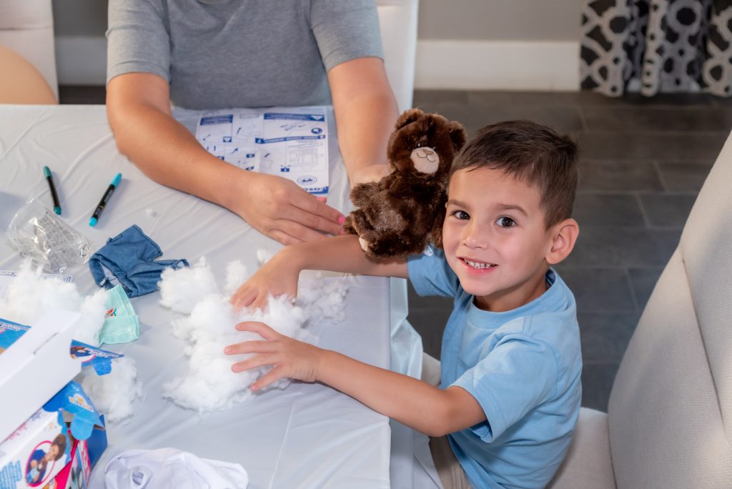 A young boy smiles while making a stuffed bear at a table covered with craft supplies and white stuffing, with an adult sitting next to him.