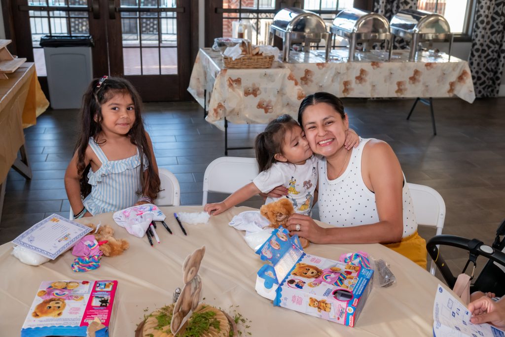 A smiling woman sits at a table with two young girls, one hugging and kissing her. Art supplies and toys are on the table. Buffet trays are set up in the background.