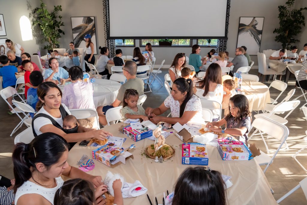 Children and adults sit at round tables in a bright room, assembling craft kits. The tables have beige cloths and decorative centerpieces. Large windows and a projector screen are visible in the background.