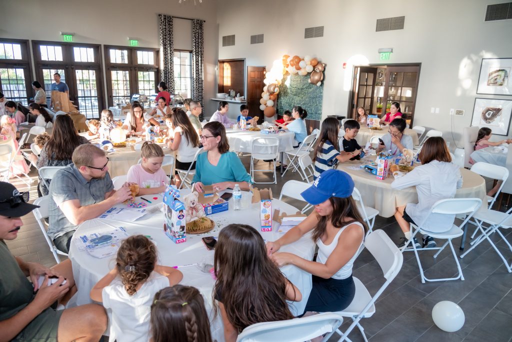 A group of people, including children and adults, sit at round tables in a bright event hall, socializing and doing activities. Balloons decorate one wall, and sunlight streams through large windows.