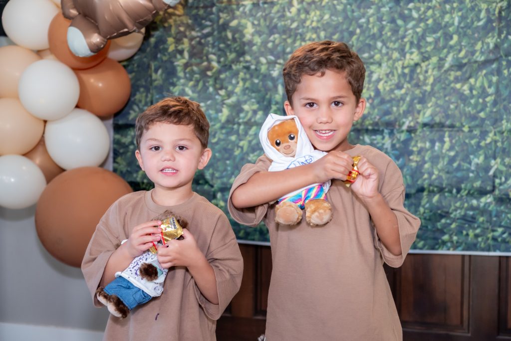 Two young boys in brown shirts smile and hold teddy bears at a party. Balloon decorations and a leafy backdrop are in the background. Both boys appear happy and are looking at the camera.