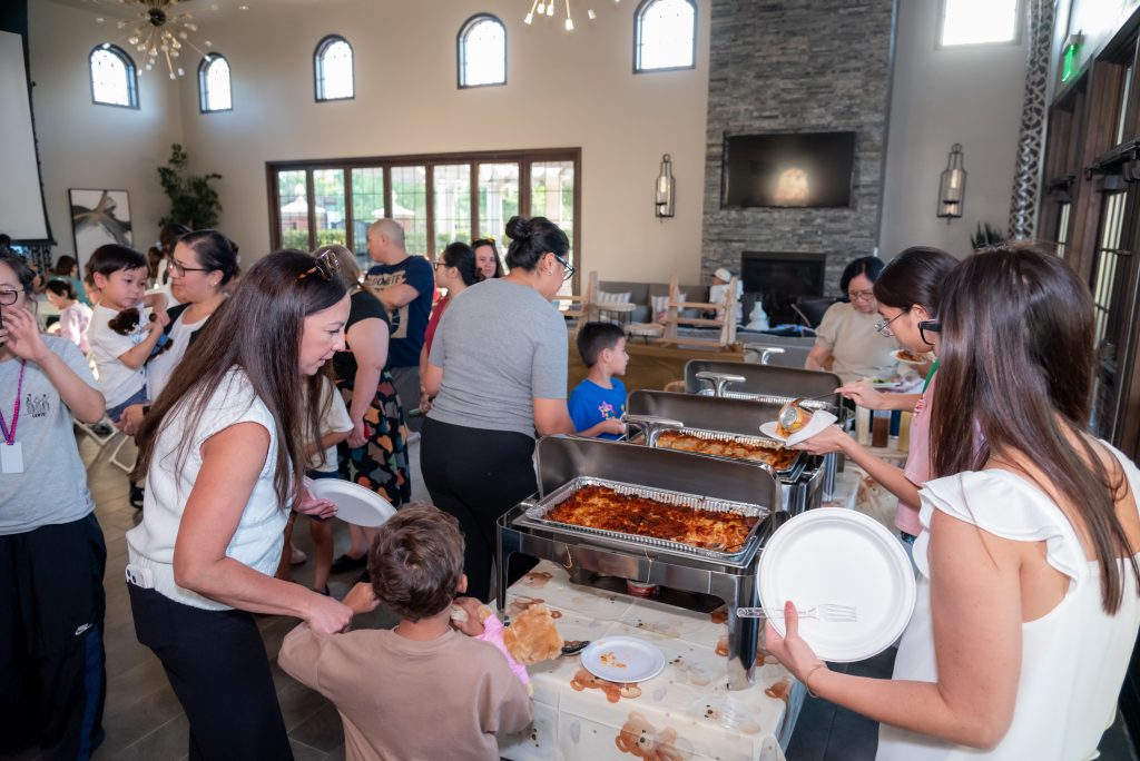 People gather around a buffet table serving food at an indoor event. Adults and children are holding plates and serving themselves, creating a lively, social atmosphere.