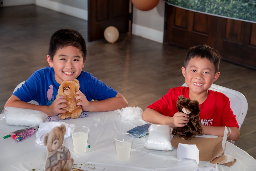 Two smiling boys sit at a table making stuffed animals. They each hold a teddy bear and have stuffing and craft supplies in front of them. The table is covered in a white cloth with drinks and teddy bear decorations.