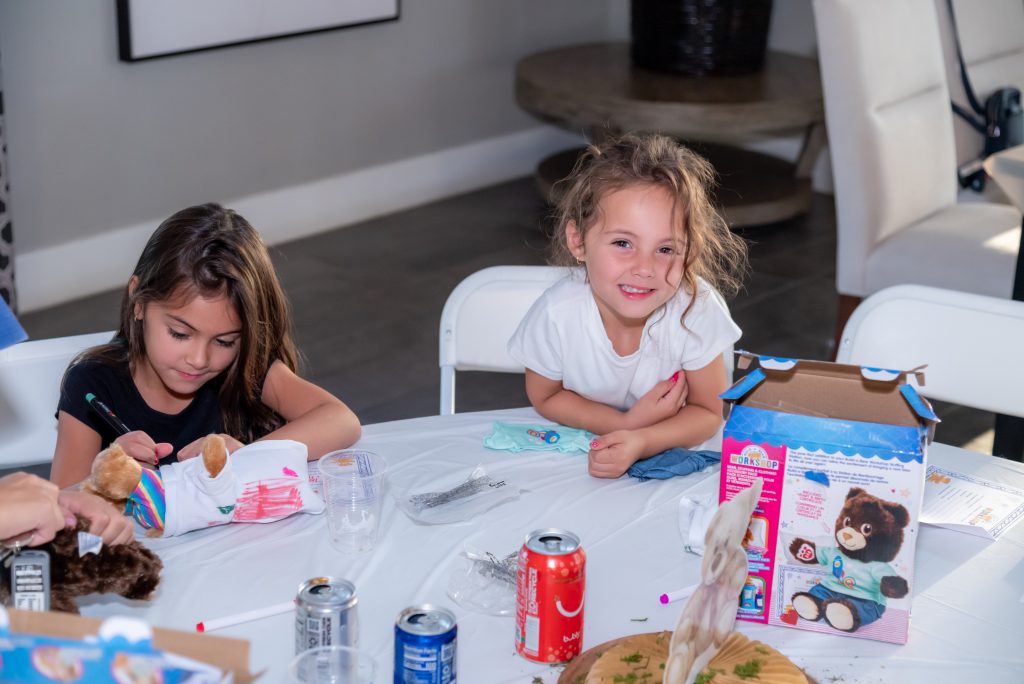 Two young girls sit at a table with soda cans, craft supplies, and stuffed animal kits. One girl draws on a stuffed bear while the other smiles happily at the camera.