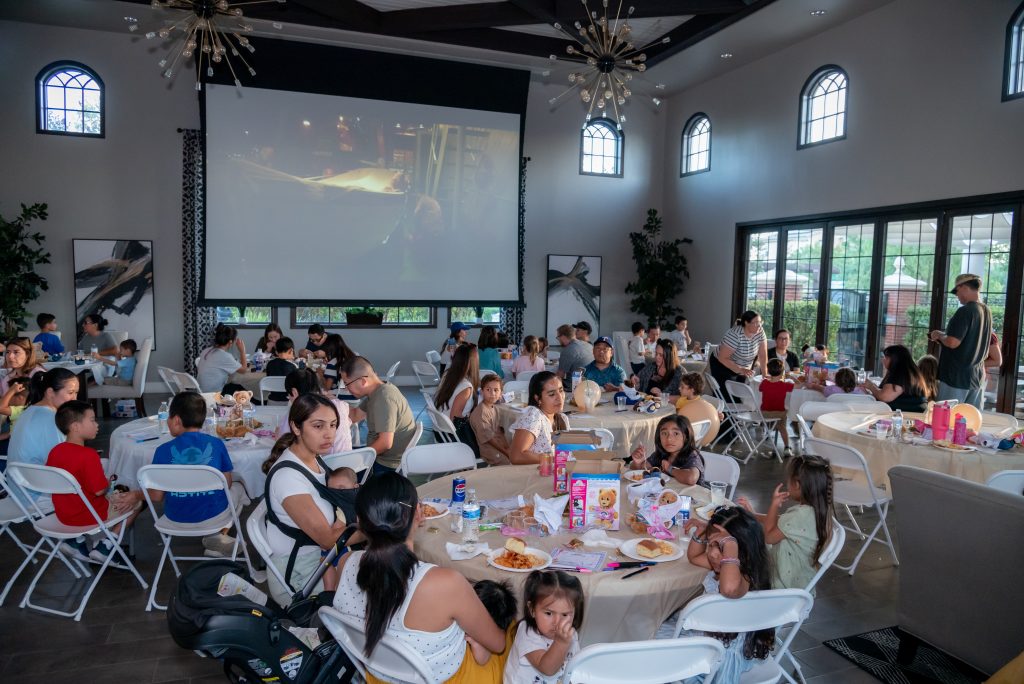 A large group of people, including many children, sit at round tables eating and watching a movie projected on a big screen in a bright, spacious room with large windows.
