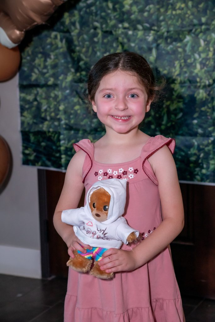 Young girl in a pink dress smiles while holding a teddy bear dressed in a white hoodie and striped shorts. There is a green leafy backdrop and brown balloons partially visible behind her.